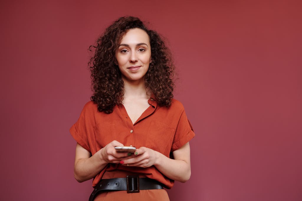 Confident businesswoman with curly hair holding a smartphone in front of a terracotta wall.